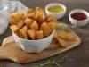 Fotografia em tons de amarelo em uma bancada de madeira escura, uma tábua de madeira, um pote branco fundo com várias mandiocas fritas. Ao fundo, um pano bege xadrez e dois potinhos um com ketchup e um de mostarda.