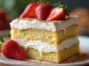 Em cima de uma mesa, prato branco com pedaço de bolo com massa branca, creme branco, recheio de creme branco, decorados com morangos.