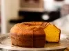 Um bolo de cenoura redondo com furo no meio está parcialmente cortado e exposto sobre um prato de metal. A textura está fofa e dourada por dentro, com uma casquinha crocante por fora.