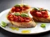 Fotografia em tons claros de três fatias de pão italiano torrado sobre um prato raso branco, apoiado em uma mesa preta. Sobre as torradas tem um pouco de confit de tomates cereja com folhas de manjericão e azeite.