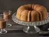 Imagem de um bolo formigueiro inteiro redondo com furo central sobre uma boleira transparente e ao lado dois copos de chocolate quente