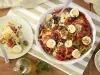 Imagem vista de cima de uma travessa redonda com bacalhoada com rodelas de ovos, pimentões, azeitonas e tomate. Ao lado, um prato branco com uma porção de bacalhoada, um vidro de azeite e um bowl de arroz branco.