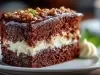 Fatia de bolo de chocolate em um prato branco. Com duas camadas de bolo úmido e um recheio cremoso, é coberto com ganache de chocolate e decorado com nozes picadas e folhas de hortelã.