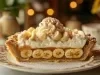 Em cima de uma mesa, prato com metade de uma banoffee com massa de biscoitos crocante, doce de leite, bananas em rodelas, decorado com chantilly cremoso, banana em rodelas e cacau em pó.