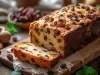 Fotografia de um bolo de frutas em uma tábua de madeira, com crosta dourada e recheio de passas. A cena inclui utensílios de cozinha e uma tigela com frutas secas.