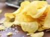 Fotografia de alguns chips de batatas sobrepostos sobre uma mesa de madeira escura, juntamente com um pouco de sal grosso. No fundo tem um saquinho verde.