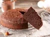 Imagem de um bolo redondo de chocolate com uma fatia em destaque. Compondo a imagem, um fuet, ovos e um pote com chocolate em pó.