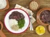 Fotografia em tons de marrom e vermelho de uma bancada de madeira, sobre ela recipientes redondos com feijoada, arroz, couve e farofa. Ao centro um paninho branco xadrez com um prato branco redondo com um prato feito de feijoada.