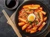Fotografia vista de cima de um recipiente fundo preto com uma receita de Tteokbokki com cebolinha e metade de um ovo cozido por cima. Ao lado tem três hashis de madeira sobre um paninho de tom bege, apoiado em uma mesa preta com texturas.