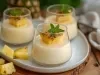 Fotografia de três copos de sobremesa com um creme claro, decorados com pedaços de abacaxi e hortelã. Os copos estão dispostos em um prato oval branco, com pedaços de abacaxi ao redor, em um ambiente acolhedor.
