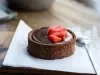 Fotografia em tons claros de uma mini torta de bombom de morango com base de chocolate sobre um papel dobrado e uma colher de sopa. Decorando a ganache, que está cobrindo a torta, tem alguns morangos decorando.