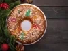 Fotografia de um bolo de reis enfeitado com frutas cristalizadas em vermelho, verde e laranja, polvilhado com açúcar e amêndoas. Colocado em um prato metálico, com ramos de pinheiro e enfeites vermelhos ao fundo.
