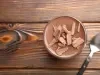 Fotografia em tons claros, vista de cima, de um recipiente de vidro pequeno com a sobremesa, finalizada com ganache e raspas de chocolate sobre uma mesa de madeira. Ao lado tem uma colher de sopa.