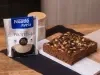 receita de bolo proteico molhadinho de banana e chocolate servida em uma porção grande sobre uma mesa de madeira decorada com um pote e uma embalagem de aveia proteína