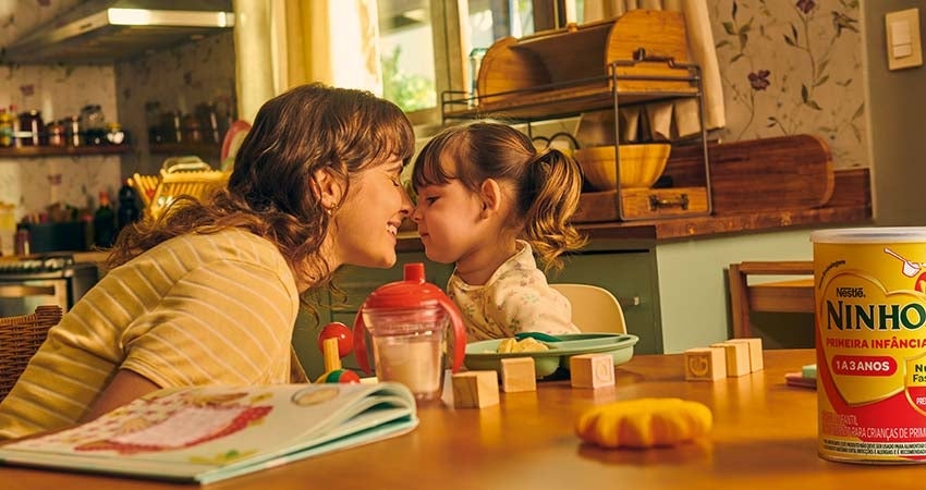 Imagem de uma mãe sorrindo bem perto de sua filha sentadas na mesa de uma cozinha. Em cima da mesa, uma lata de Ninho, alguns
