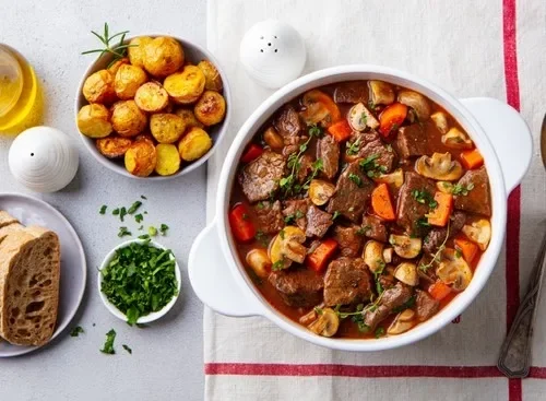Ensopado de carne com legumes em tigela branca, acompanhado de batatas assadas, pão fatiado e ervas frescas na mesa.