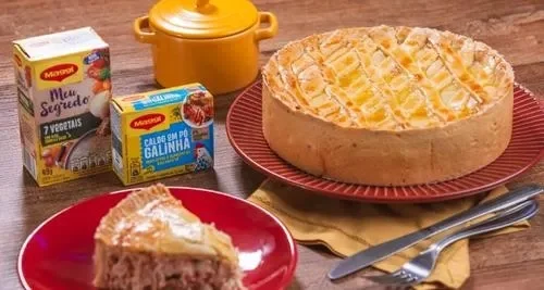 Torta salgada dourada em prato vermelho com fatia servida ao lado, acompanhada de temperos Maggi e panela amarela.