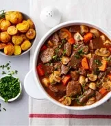 Ensopado de carne com legumes em tigela branca, acompanhado de batatas assadas, pão fatiado e ervas frescas na mesa.