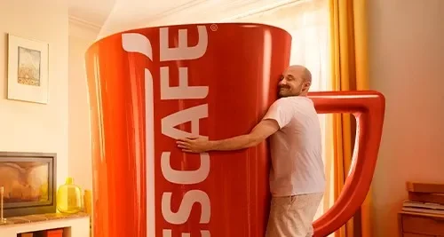 Homem abraça uma xícara gigante NESCAFÉ em sala iluminada, com decoração aconchegante em tons quentes.