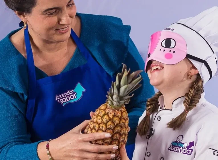 Responsável e criança caracterizada como chef seguram abacaxi com venda nos olhos, em atividade culinária com Ascenda.